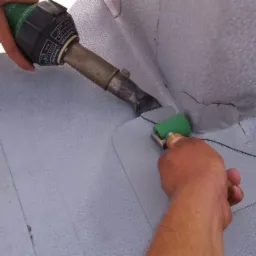 Prace dekarskie: Zgrzewanie membrany dachowej za pomocą zgrzewarki i wałka dociskowego, widoczne łączenie arkuszy.