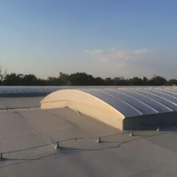 Szary dach z dwoma świetlikami łukowymi, widoczny maszt antenowy w tle, błękitne niebo.