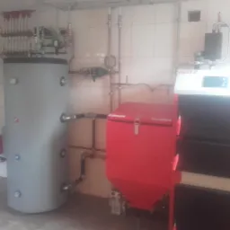Kotłownia z kotłem na paliwo stałe marki Heitz, zasobnikiem wody i rozdzielaczem, widoczne rury instalacji grzewczej na tle białych płytek.