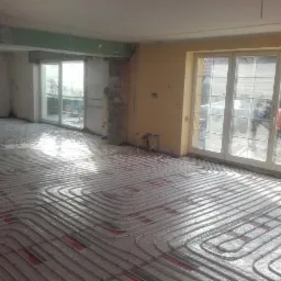 Białe rury ogrzewania podłogowego ułożone na siatce w jasnym, surowym wnętrzu. Widoczne okna balkonowe i fragmenty ścian w trakcie wykończenia.