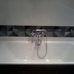 Biała wanna z chromowaną baterią i geometrycznym wzorem płytek w odcieniach szarości. Nowoczesne wykończenie łazienki z subtelnym oświetleniem.