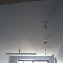 Gładki, pomalowany sufit z równo rozmieszczonymi punktami oświetleniowymi. Ściana z delikatnym wzorem w tle. Widok z dołu, wnętrze po remoncie.