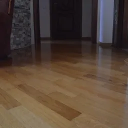 Błyszcząca, parkietowa podłoga z drewna dębowego, ułożona w jodełkę, z widocznym fragmentem brązowego fotela i kamiennej ściany w tle.