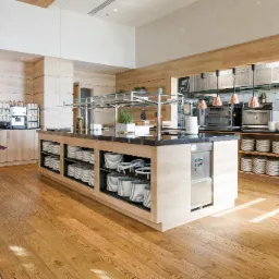 Jasna podłoga drewniana w restauracji z wyspą bufetową, na której znajdują się stosy białych talerzy, z widocznymi maszynami do kawy i piecami w tle.
