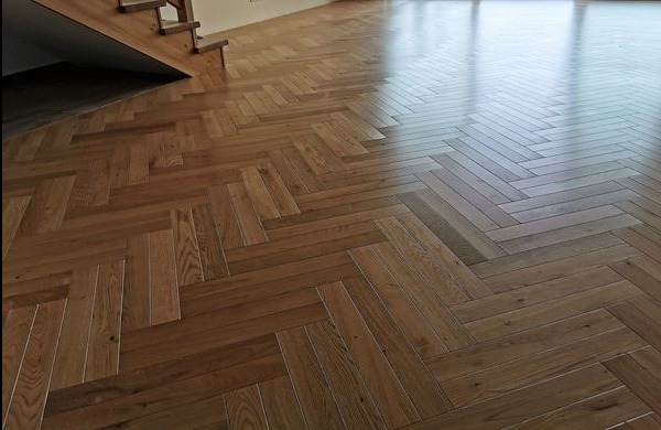 Parkiet ułożony w jodełkę francuską, wykonany z lakierowanego drewna dębowego, z widocznymi schodami w tle i odbiciem światła w polerowanej powierzchni.