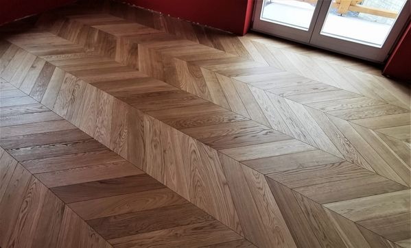 Parkiet ułożony we wzór jodełki francuskiej, wykonany z desek o naturalnym odcieniu drewna, widoczny fragment czerwonej ściany i drzwi balkonowe.