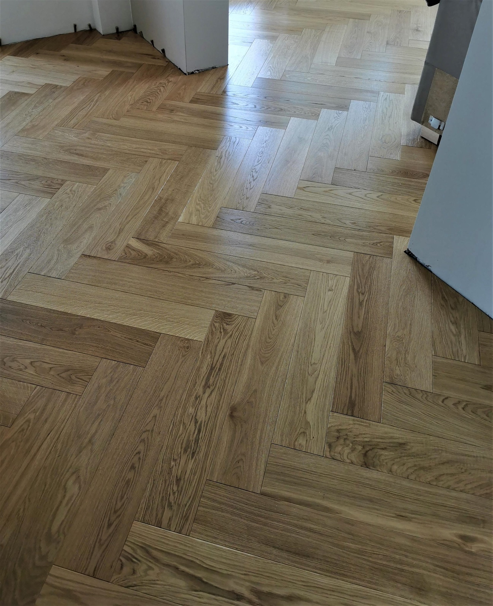 Ułożona w jodełkę jasna podłoga z desek drewnianych, widoczne szczeliny dylatacyjne przy ścianie, fragment futryny drzwi.