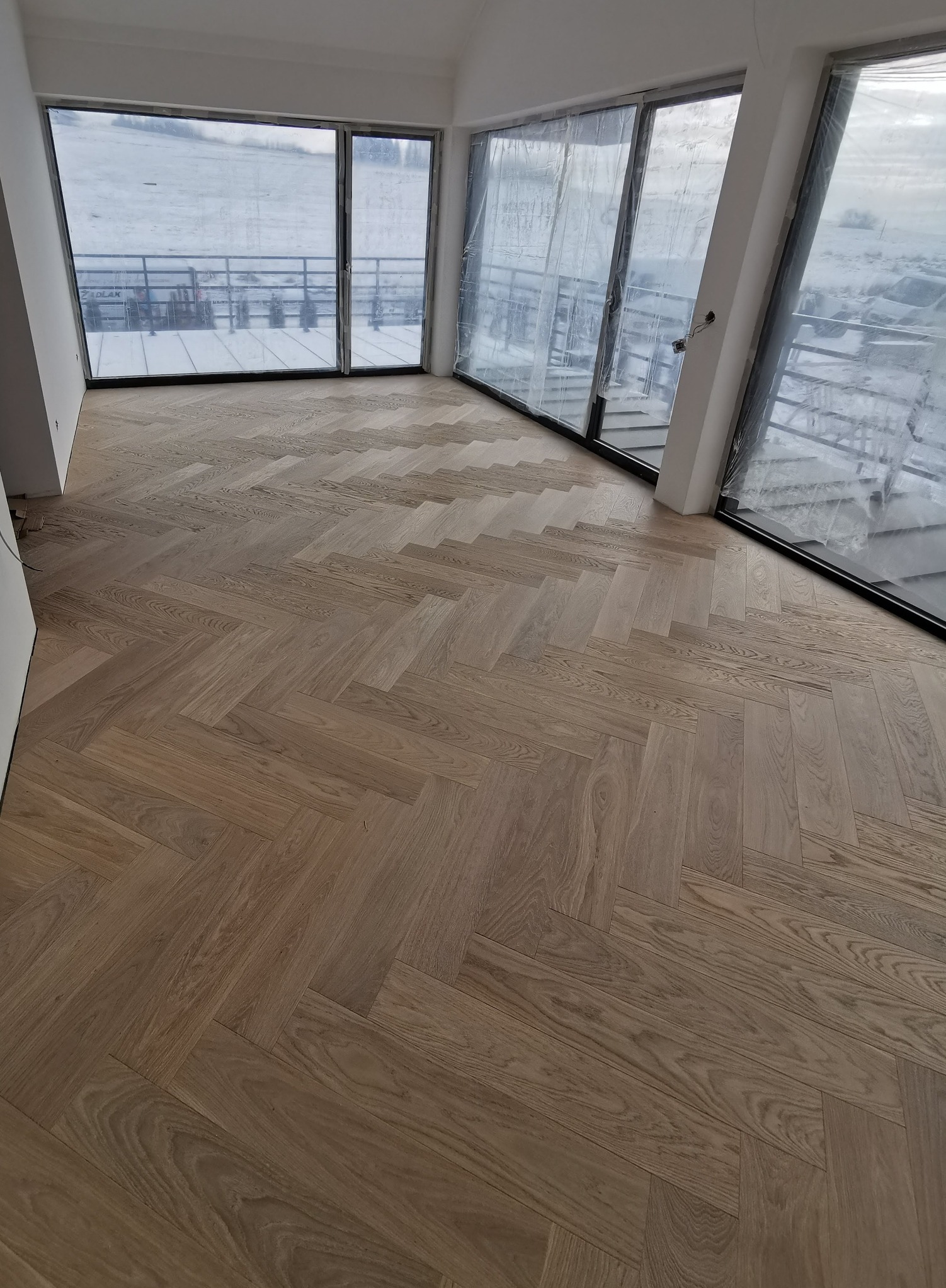 Nowa, jasna podłoga drewniana ułożona w jodełkę francuską w przestronnym pokoju z dużymi oknami, za którymi widać zimowy krajobraz.