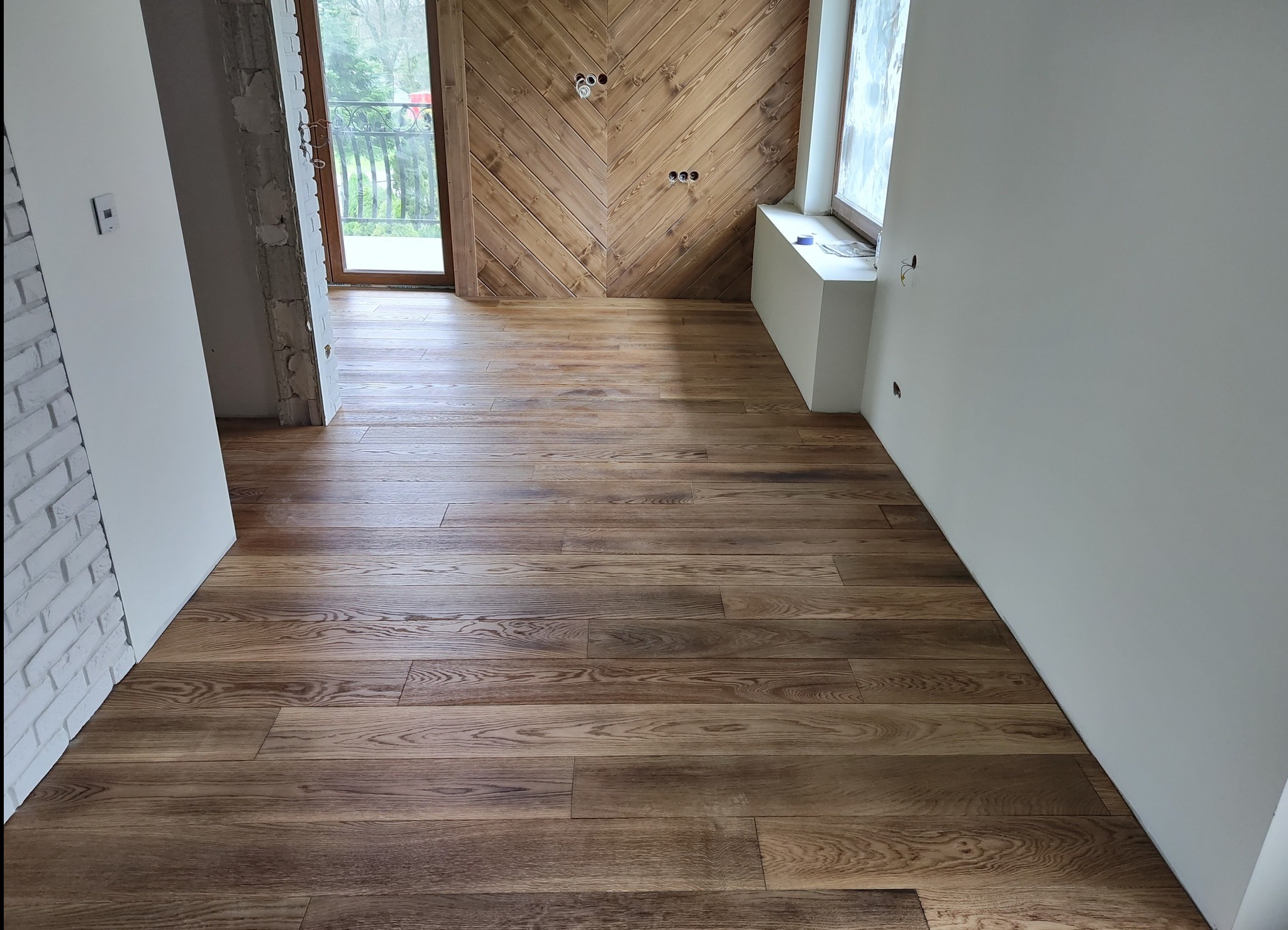 Wnętrze w trakcie wykańczania: świeżo położona, lakierowana podłoga z desek w odcieniach brązu, ściana z drewnianą boazerią ułożoną w jodełkę, widoczne puszki elektryczne bez osprzętu, surowa...