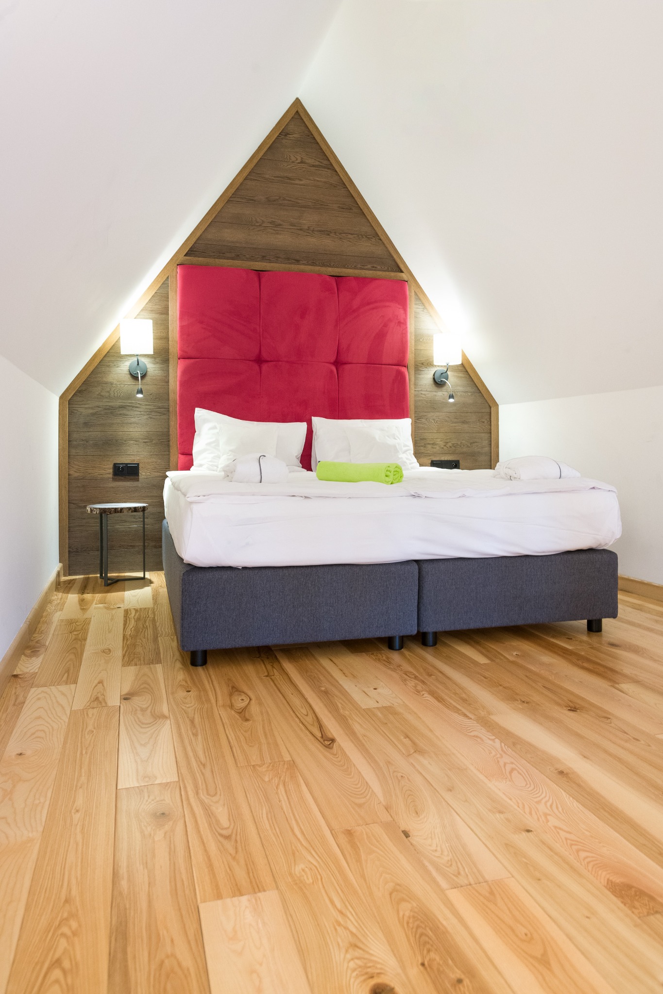 Jasna drewniana podłoga w sypialni na poddaszu z łóżkiem z czerwonym zagłówkiem i białymi poduszkami, widoczne lampki nocne i stolik.