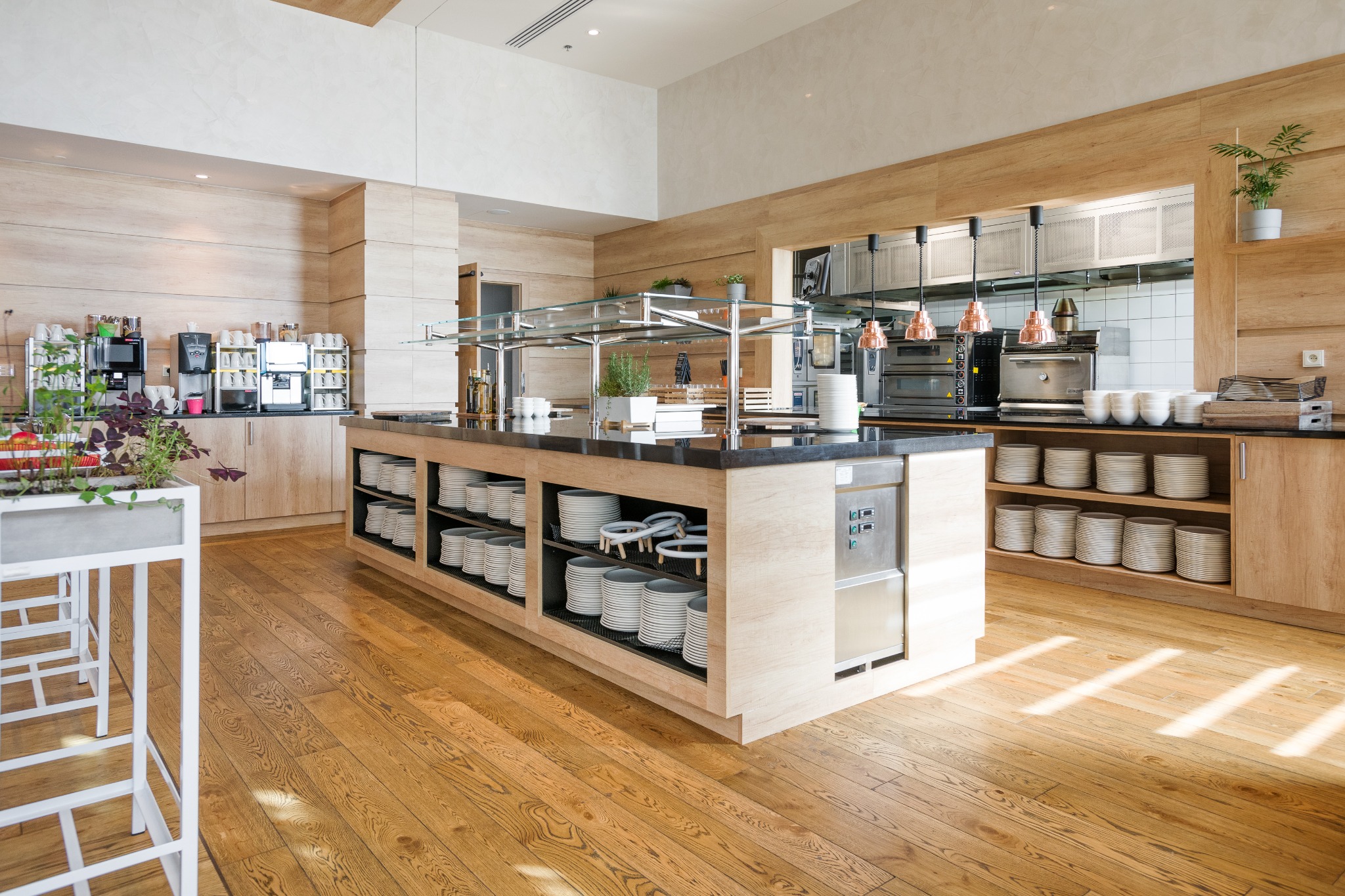 Jasna podłoga drewniana w restauracji z wyspą bufetową, na której znajdują się stosy białych talerzy, z widocznymi maszynami do kawy i piecami w tle.