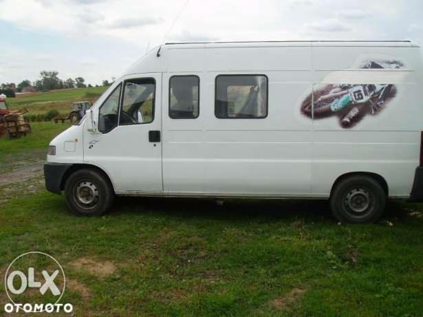 Biały bus z grafiką motocykla crossowego na boku, zaparkowany na trawiastym terenie. Widoczne okna i koła, w tle pole i zabudowania gospodarcze.