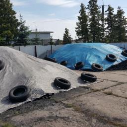 Składy węgla Pawłowski - Składowisko węgla: dwa kopce przykryte plandekami (szara i niebieska), obciążone oponami na betonowym placu. W tle budynek i drzewa.