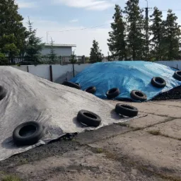 Składowisko węgla: dwa kopce przykryte plandekami (szara i niebieska), obciążone oponami na betonowym placu. W tle budynek i drzewa.