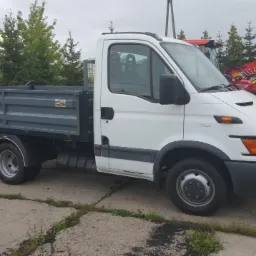 Biały samochód dostawczy z szarą skrzynią ładunkową na tle zieleni i czerwonej koparki. Widoczne opony i maszt telekomunikacyjny w tle.