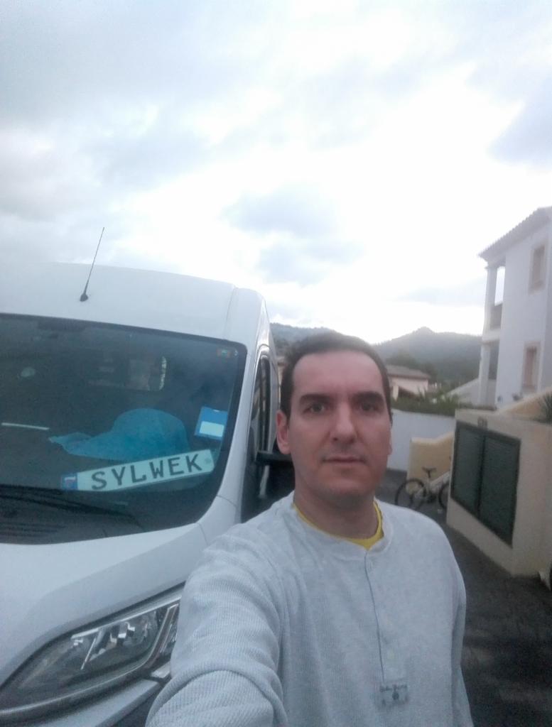 Mężczyzna robi selfie na tle białego busa z tablicą rejestracyjną 'SYLWEK' w Zabierzowie, w tle budynek mieszkalny i pochmurne niebo.