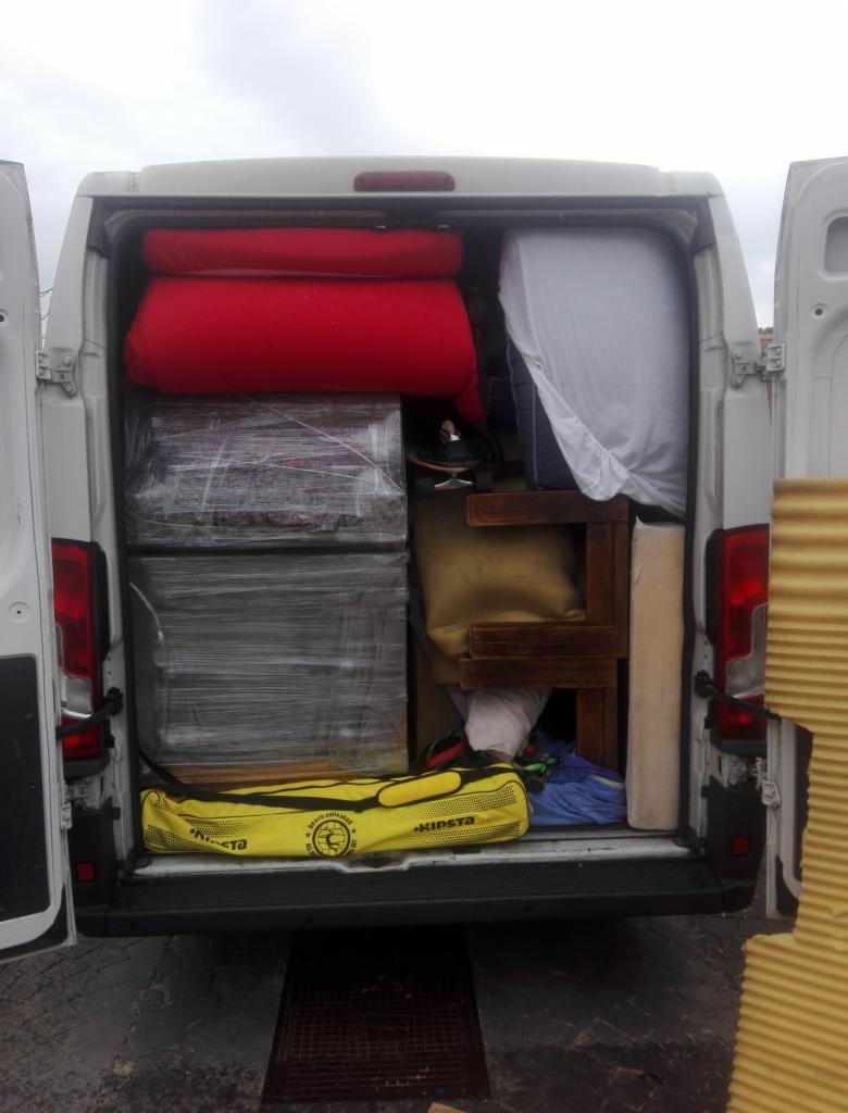 Wnętrze białego busa załadowane meblami, materacami, kartonami owiniętymi folią stretch i torbą sportową Kipsta, widok z tyłu przez otwarte drzwi.