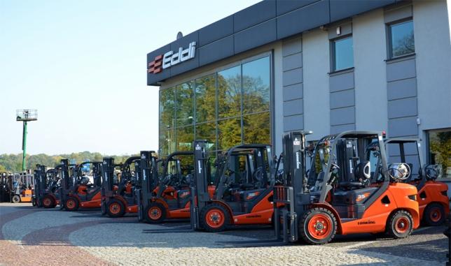 Pomarańczowe wózki widłowe marki Turning, ustawione rzędem przed budynkiem firmy Eddi, w słoneczny dzień. Widoczna wieża obserwacyjna w tle.