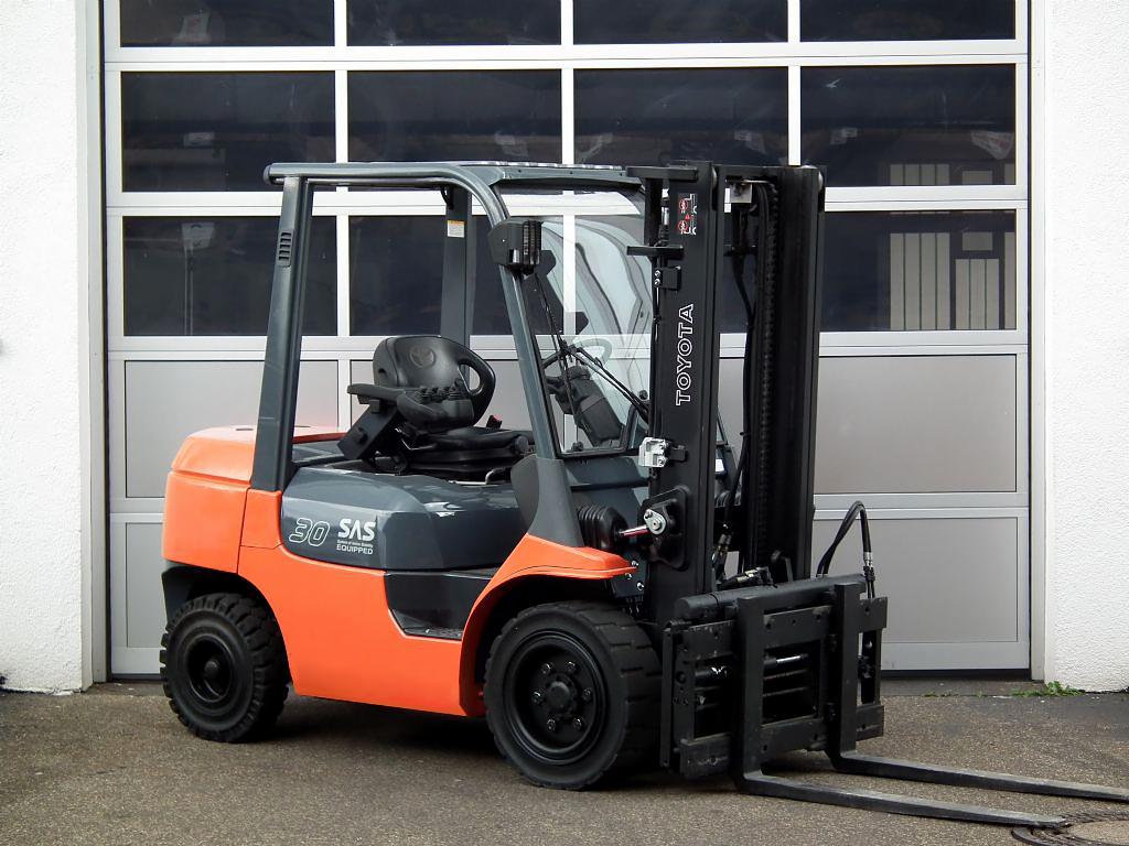 Pomarańczowo-szary wózek widłowy Toyota 30 SAS na tle białej ściany i przeszklonej bramy garażowej. Widok z boku, lekko od dołu.