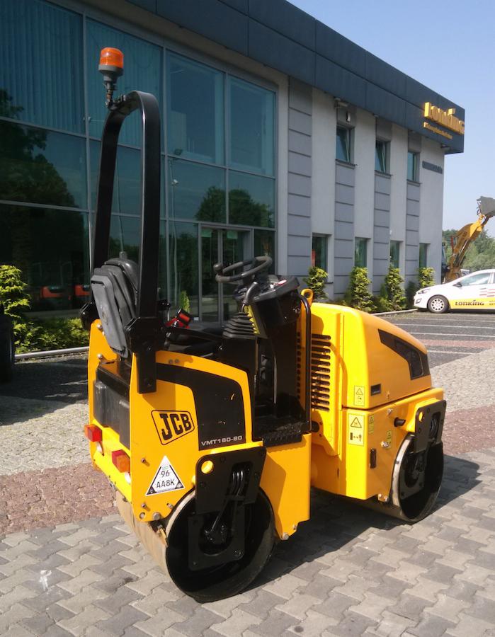 Żółty walec drogowy JCB VMT160-80 na kostce brukowej przed budynkiem z dużymi oknami. Widoczny fragment wózka widłowego i samochodu w tle.