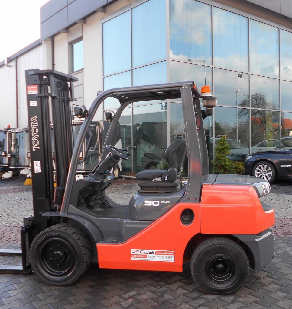 Pomarańczowo-szary wózek widłowy Toyota Geneo 30 na tle budynku z lustrzanymi oknami, z widocznymi detalami jak logo Eddi i inne pojazdy w tle.