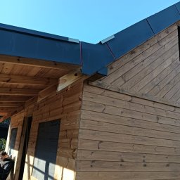 DEKER ZR SPÓŁKA Z OGRANICZONĄ ODPOWIEDZIALNOŚCIĄ - Budynek z drewnianą elewacją z poziomych i ukośnych desek, z ciemnym, panelowym pokryciem dachowym, widoczne krokwie i błękitne niebo.