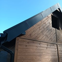 DEKER ZR SPÓŁKA Z OGRANICZONĄ ODPOWIEDZIALNOŚCIĄ - Nowoczesny budynek z ciemnym, panelowym dachem i dopasowanymi rynnami, kontrastujący z elewacją z drewnianych desek ułożonych poziomo i ukośnie na szczycie.