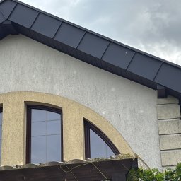 DEKER ZR SPÓŁKA Z OGRANICZONĄ ODPOWIEDZIALNOŚCIĄ - Ciemny dach z metalowych paneli na rąbek stojący, z widoczną rynną, nad jasną elewacją z trzema łukowymi oknami w brązowych ramach, pod zachmurzonym niebem.