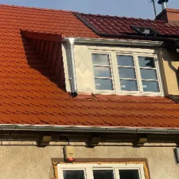 Czerwona dachówka karpiówka na dachu z oknem lukarną i obróbką blacharską wokół okna, widoczny system rynnowy i komin w tle.