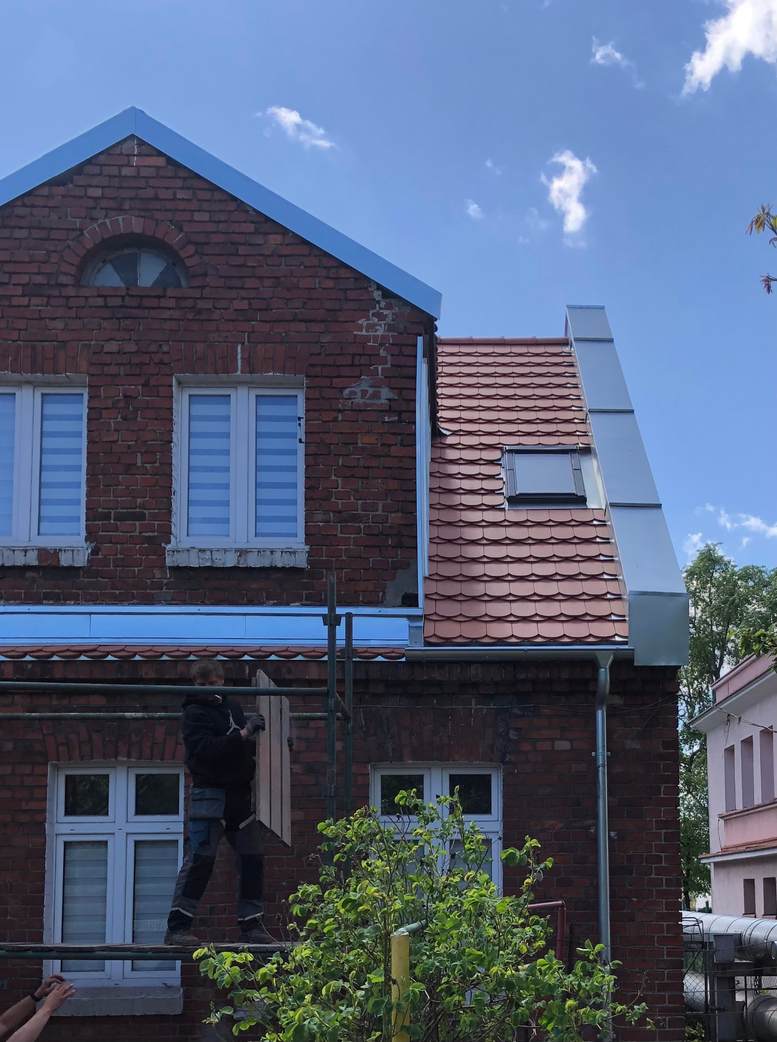 Pracownik na rusztowaniu, montujący element dachu na tle ceglanego budynku z nowym pokryciem dachowym z dachówki ceramicznej i obróbkami blacharskimi.