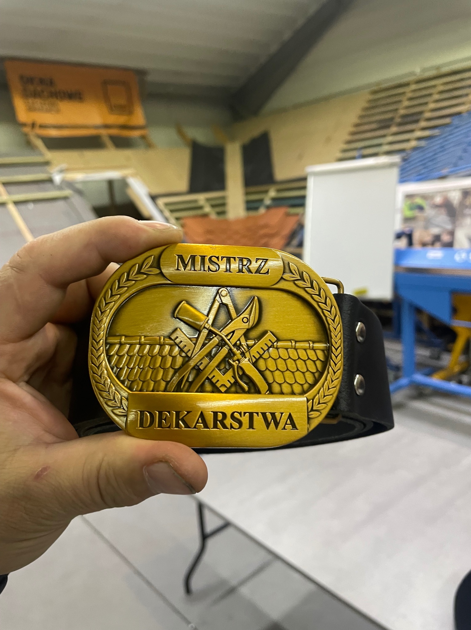 Złota odznaka na czarnym pasku z napisem 'Mistrz Dekarstwa', trzymana w dłoni, w tle widoczne elementy konstrukcji dachu i materiały budowlane.