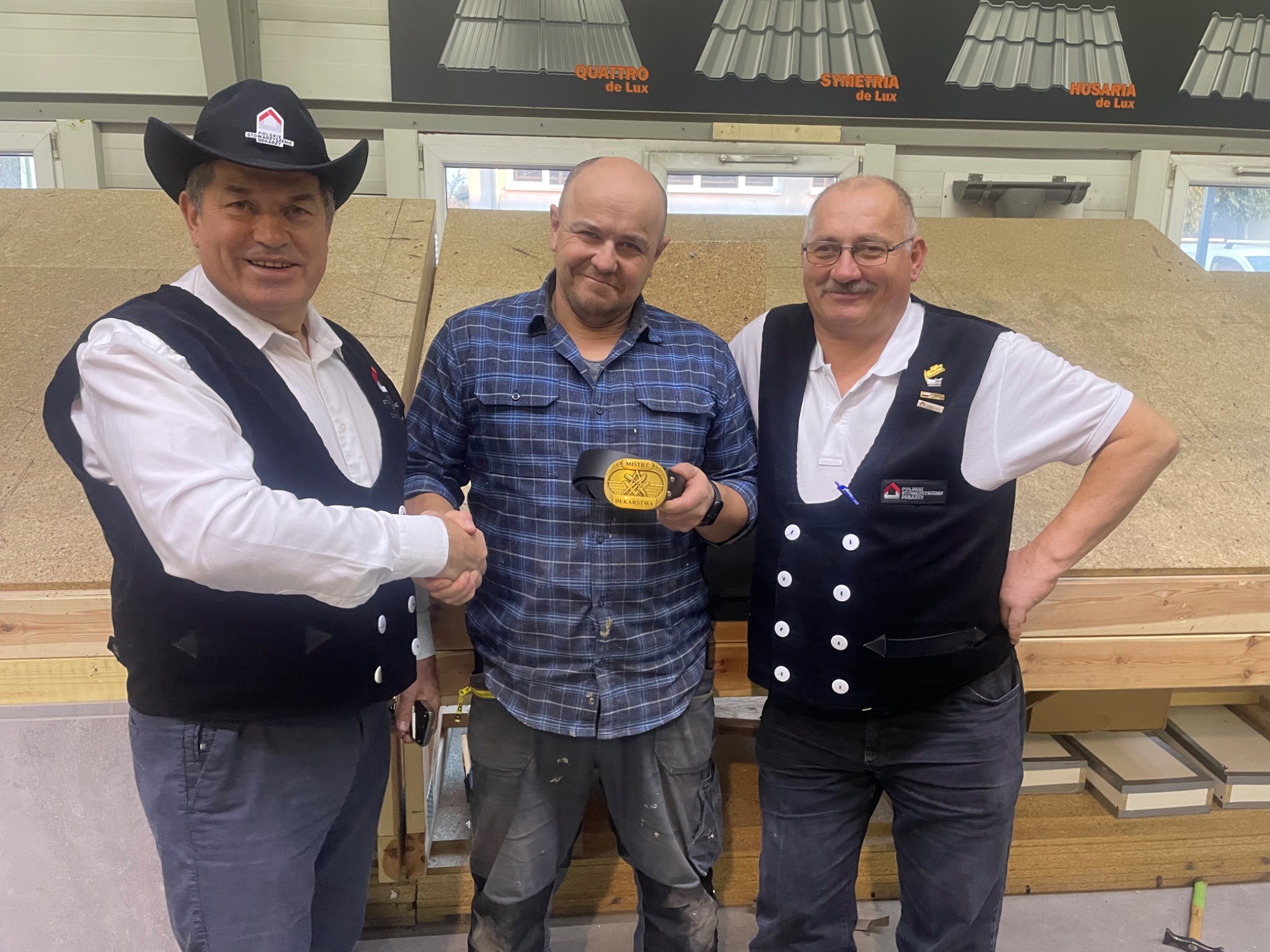 Trzech dekarzy, w tym jeden w kowbojskim kapeluszu, ściska dłoń mężczyźnie trzymającemu nagrodę 'Mistrz Dekarstwa' na tle ekspozycji różnych rodzajów pokryć dachowych.