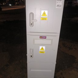P&K - Szara, dwuczęściowa skrzynka elektryczna z logo Energa i ostrzeżeniami o napięciu, umieszczona na ziemi, nocne oświetlenie w tle. Widoczne zamki i etykiety producenta.