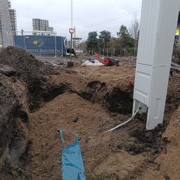 P&K - Wykop z kablem i białą skrzynką przyłączeniową na budowie, w tle budynki i ogrodzenie z logo dewelopera. Widoczna łopata i inne narzędzia.