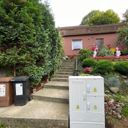 P&K - Biała skrzynka elektryczna z ostrzeżeniami na tle domu z ogrodem, widoczna ziemia po wykopie. Ujęcie z dołu, pochmurne niebo.