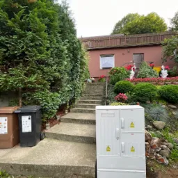 Biała skrzynka elektryczna z ostrzeżeniami na tle domu z ogrodem, widoczna ziemia po wykopie. Ujęcie z dołu, pochmurne niebo.