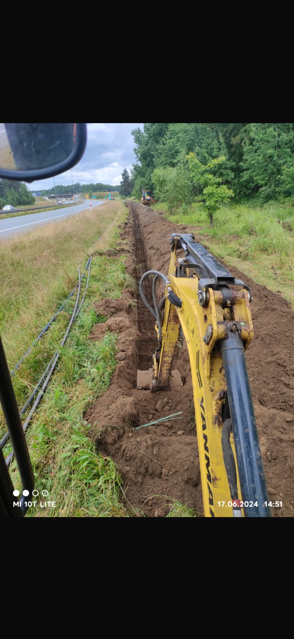 Żółta minikoparka Yanmar w trakcie wykonywania długiego, wąskiego wykopu w ziemi wzdłuż drogi, z widocznymi rurami ułożonymi obok wykopu, perspektywa z wnętrza kabiny operatora.
