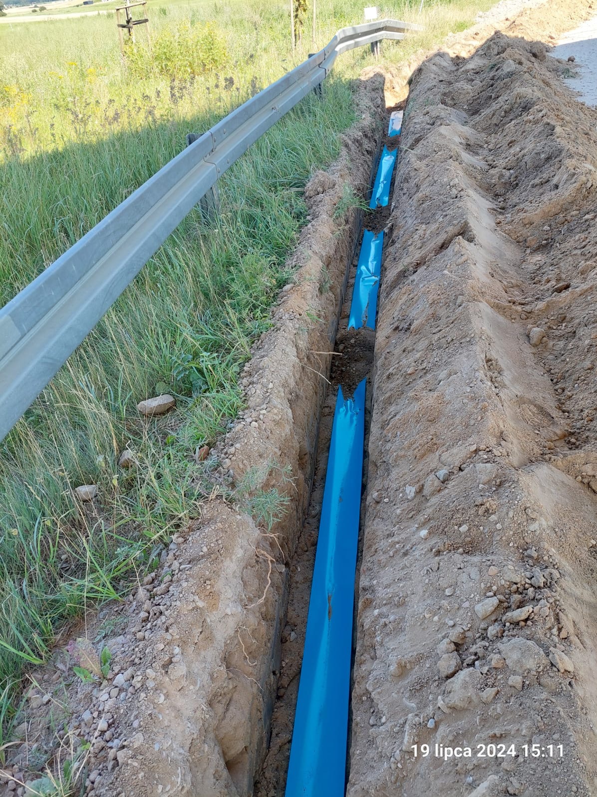 Wąski wykop w ziemi z ułożonym niebieskim rurociągiem, widoczne ślady niedawnych prac ziemnych, w tle trawa i bariera ochronna.