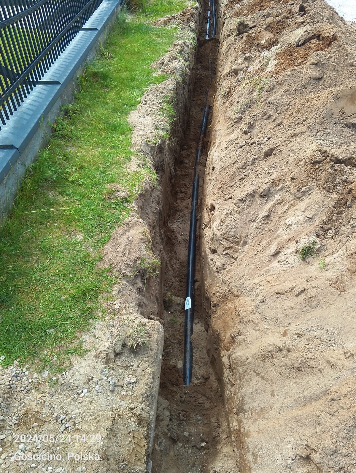 Wąski wykop w ziemi z ułożonym czarnym rurociągiem, widoczny trawnik i fragment ogrodzenia