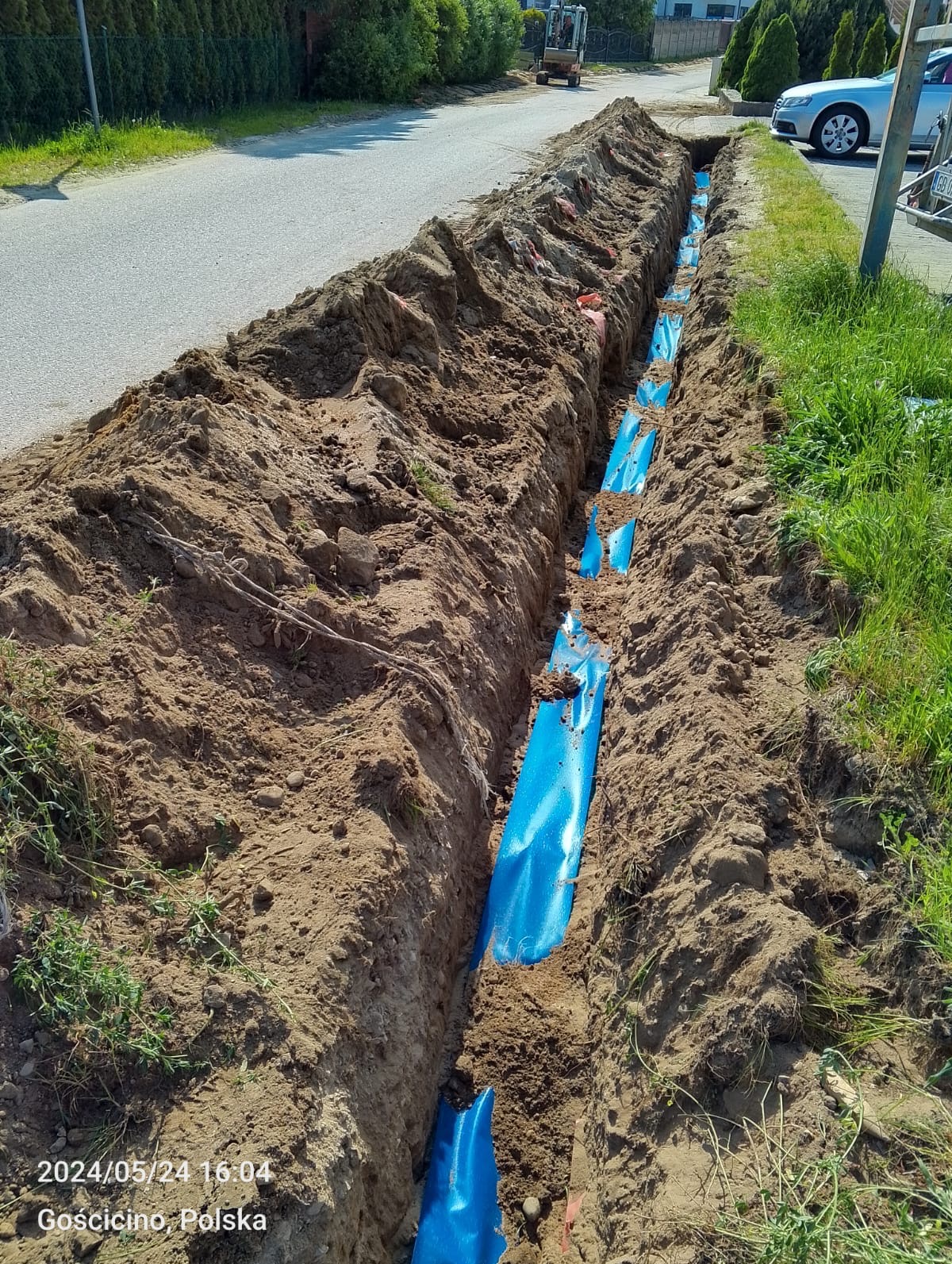 Wykopany rów z niebieską rurą instalacyjną na dnie, widok z góry na tle ulicy i zieleni.