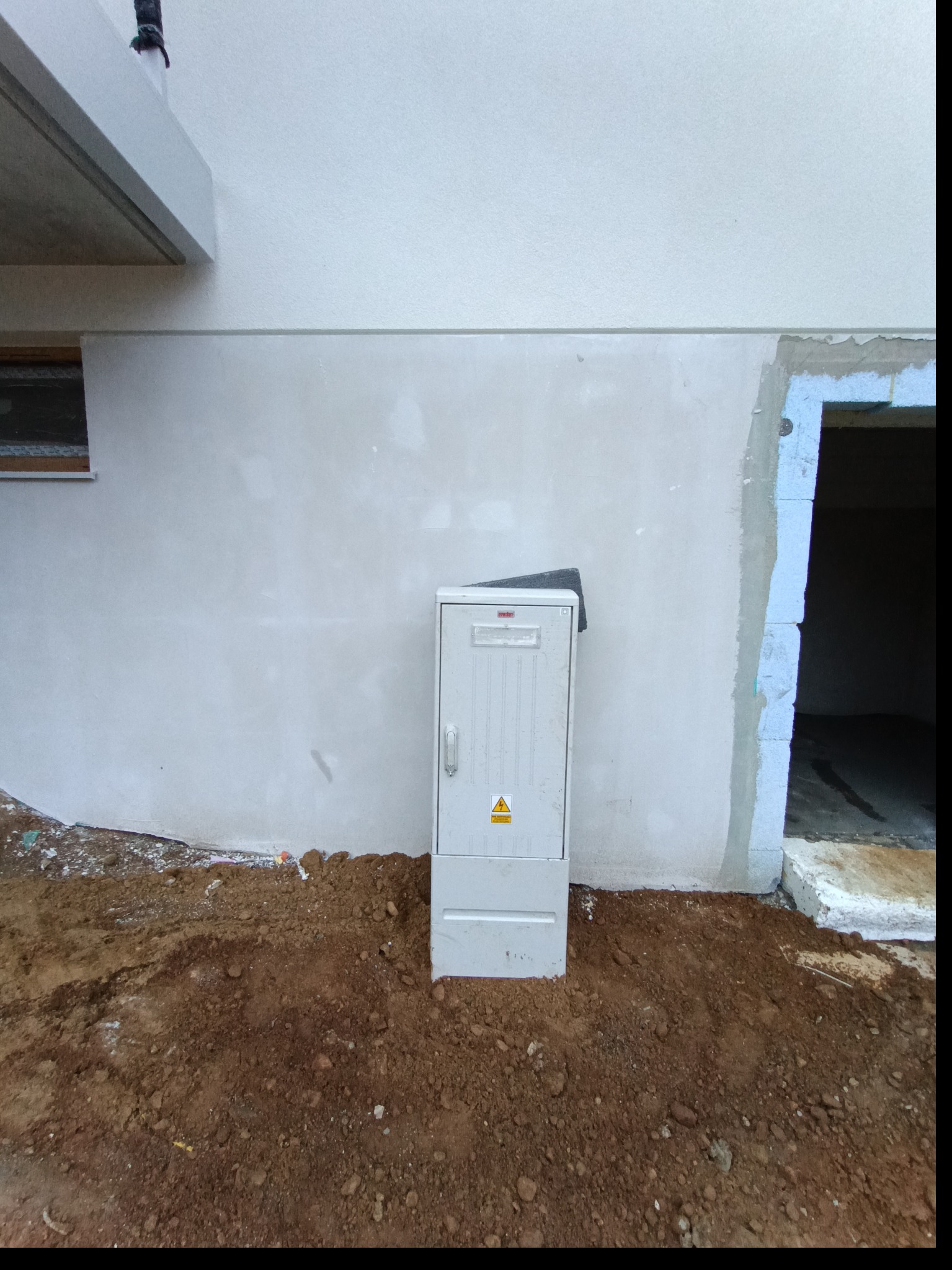 Szara skrzynka elektryczna z symbolem ostrzegawczym na tle ściany budynku w trakcie budowy, z widocznym wejściem i ziemią. Ujęcie z bliska, z dołu.