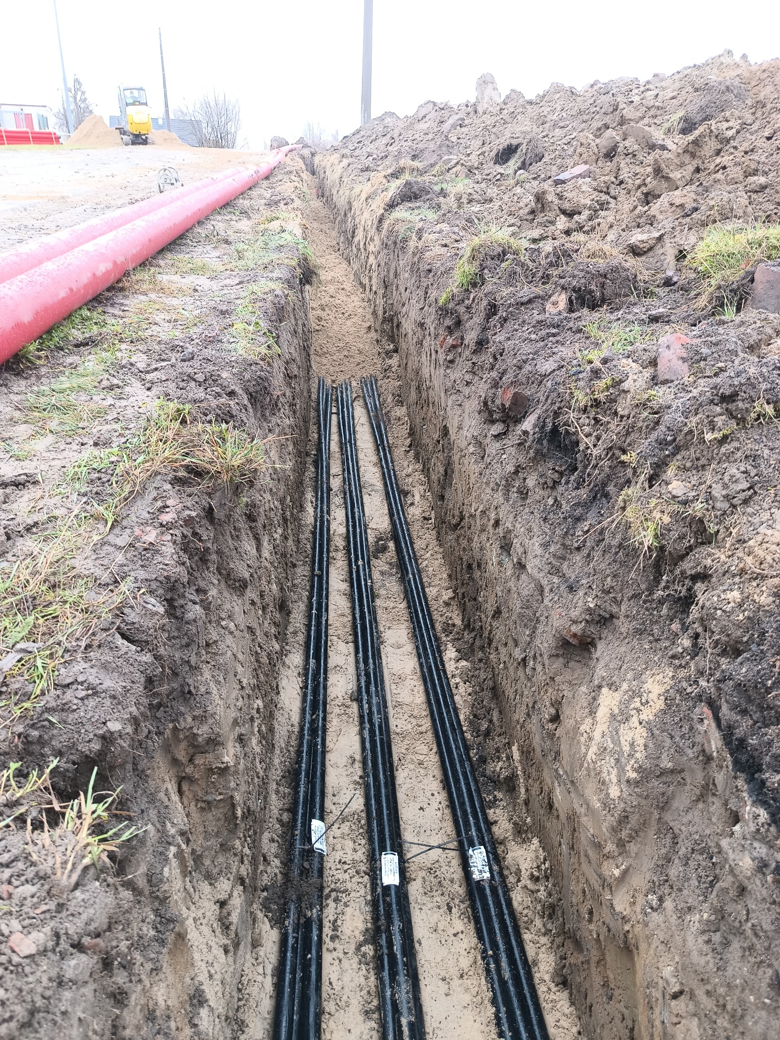 Wąski wykop z trzema czarnymi rurami na dnie, widoczne czerwone rury na powierzchni i mała koparka w tle. Grunt piaszczysto-gliniasty.