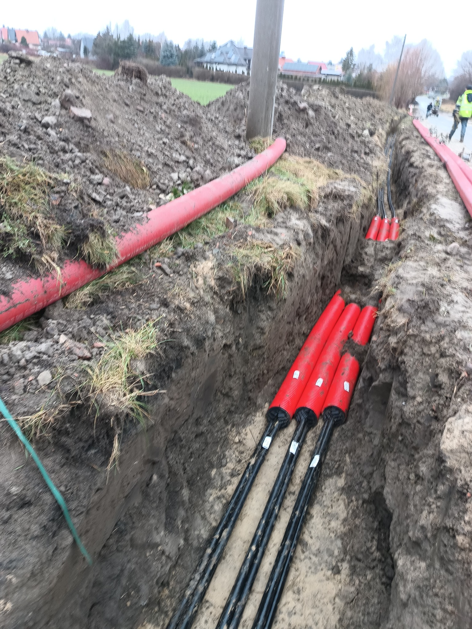 Wykop z ułożonymi czerwonymi rurami osłonowymi i kablami energetycznymi w ziemi. Widoczne zakończenia kabli z oznaczeniami. Prace ziemne w toku, widoczny krajobraz.
