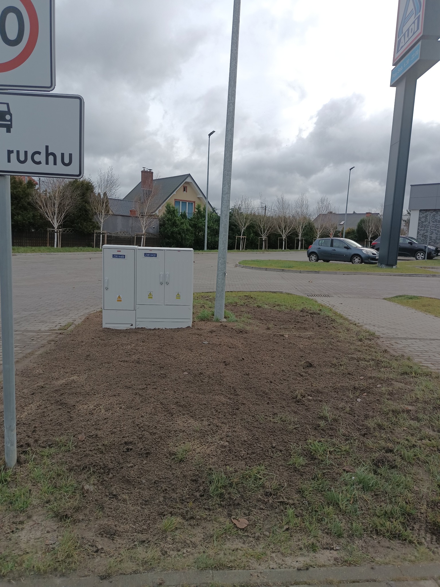 Szara szafa elektryczna z oznaczeniami ostrzegawczymi, umieszczona na ziemi obok parkingu i chodnika, z budynkiem w tle i znakiem Aldi w prawym górnym rogu.