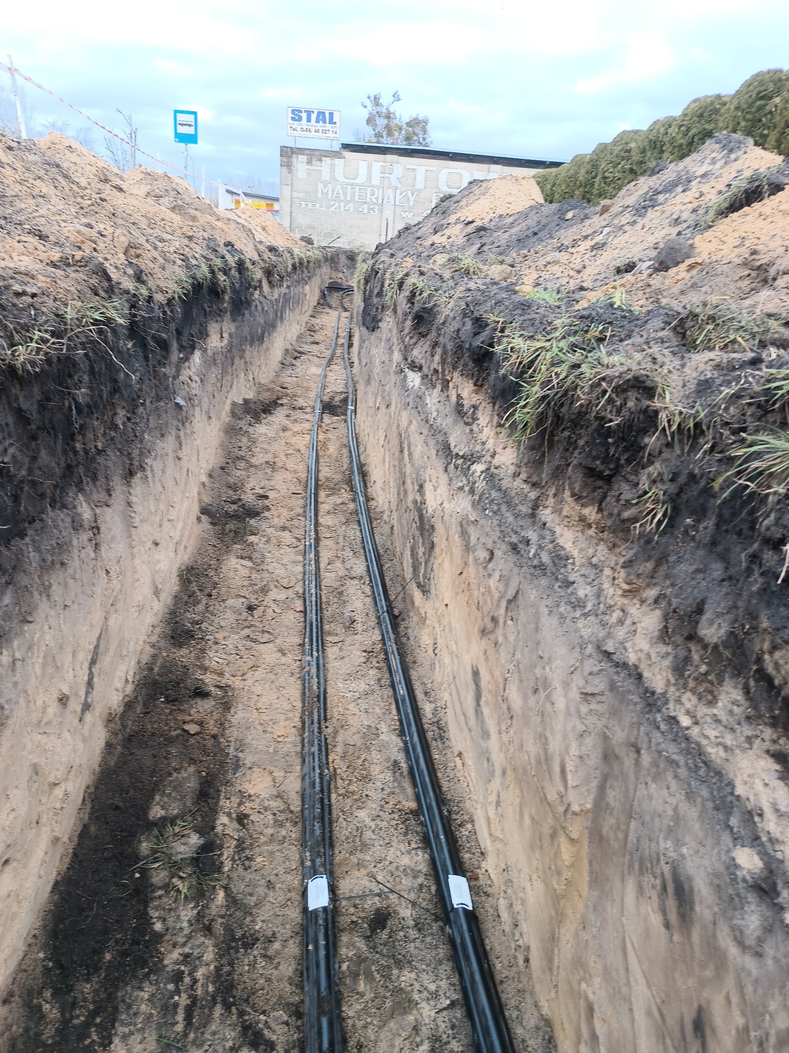 Wąski wykop z ułożonymi kablami w ziemi. Widoczne ściany wykopu z warstwami ziemi i niewielką roślinnością. W tle budynek z reklamą.