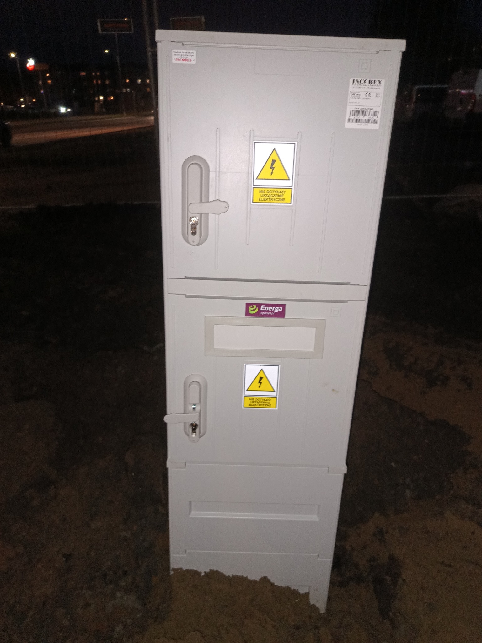 Szara, dwuczęściowa skrzynka elektryczna z logo Energa i ostrzeżeniami o napięciu, umieszczona na ziemi, nocne oświetlenie w tle. Widoczne zamki i etykiety producenta.
