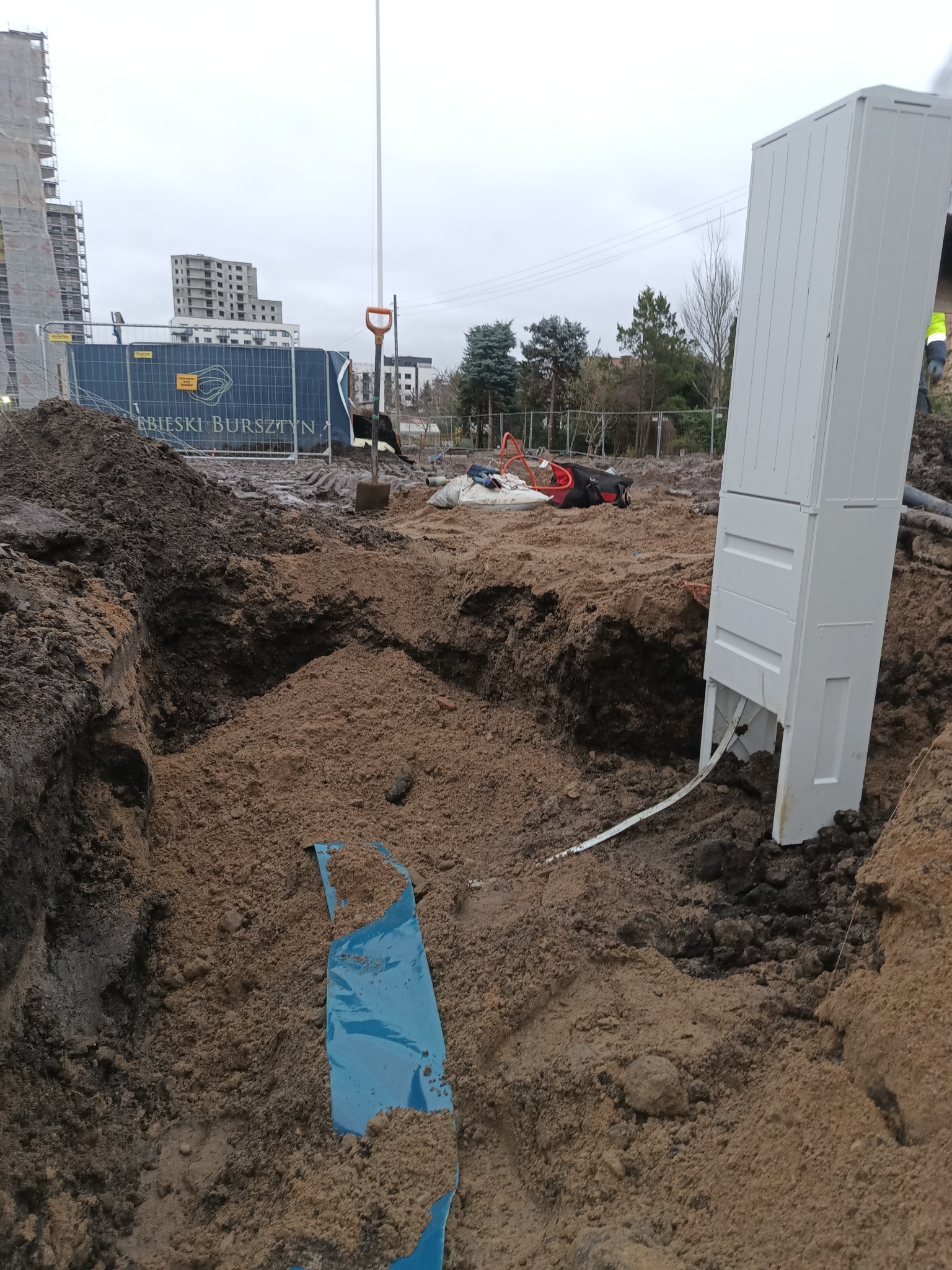 Wykop z kablem i białą skrzynką przyłączeniową na budowie, w tle budynki i ogrodzenie z logo dewelopera. Widoczna łopata i inne narzędzia.