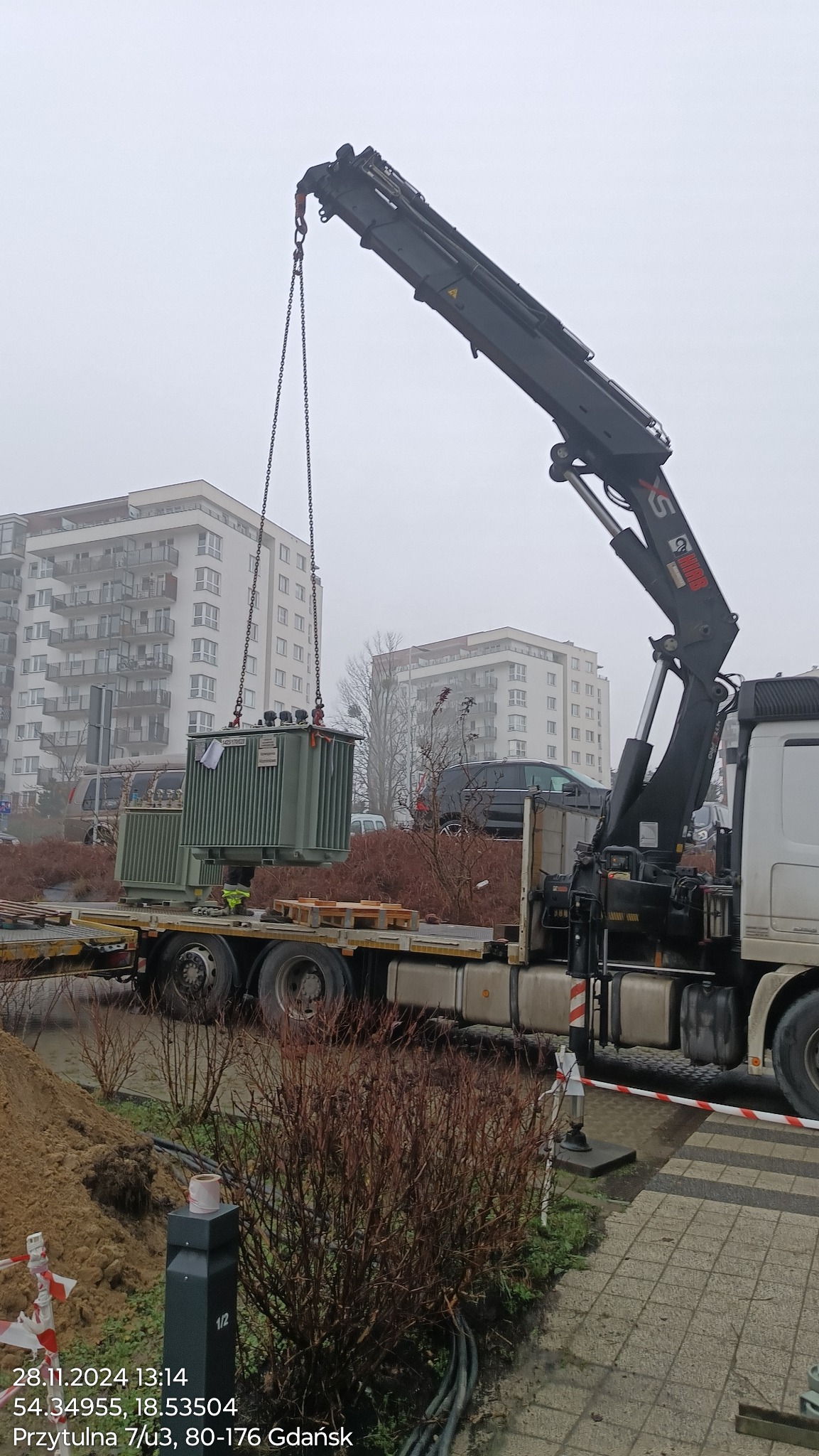 Dźwig samochodowy podnosi transformator na tle miejskiego krajobrazu w pochmurny dzień. Widoczne budynki mieszkalne i elementy infrastruktury.