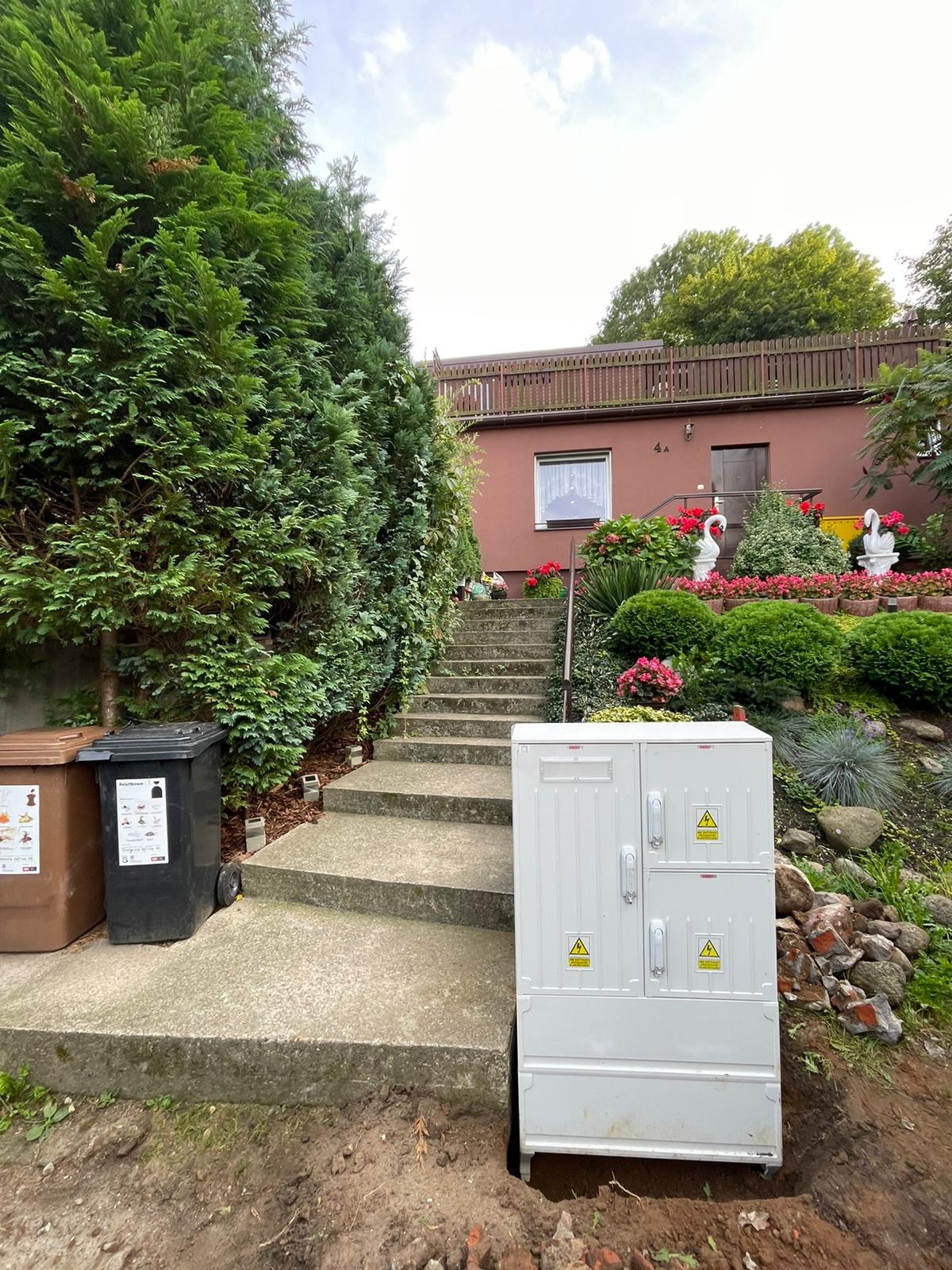 Biała skrzynka elektryczna z ostrzeżeniami na tle domu z ogrodem, widoczna ziemia po wykopie. Ujęcie z dołu, pochmurne niebo.