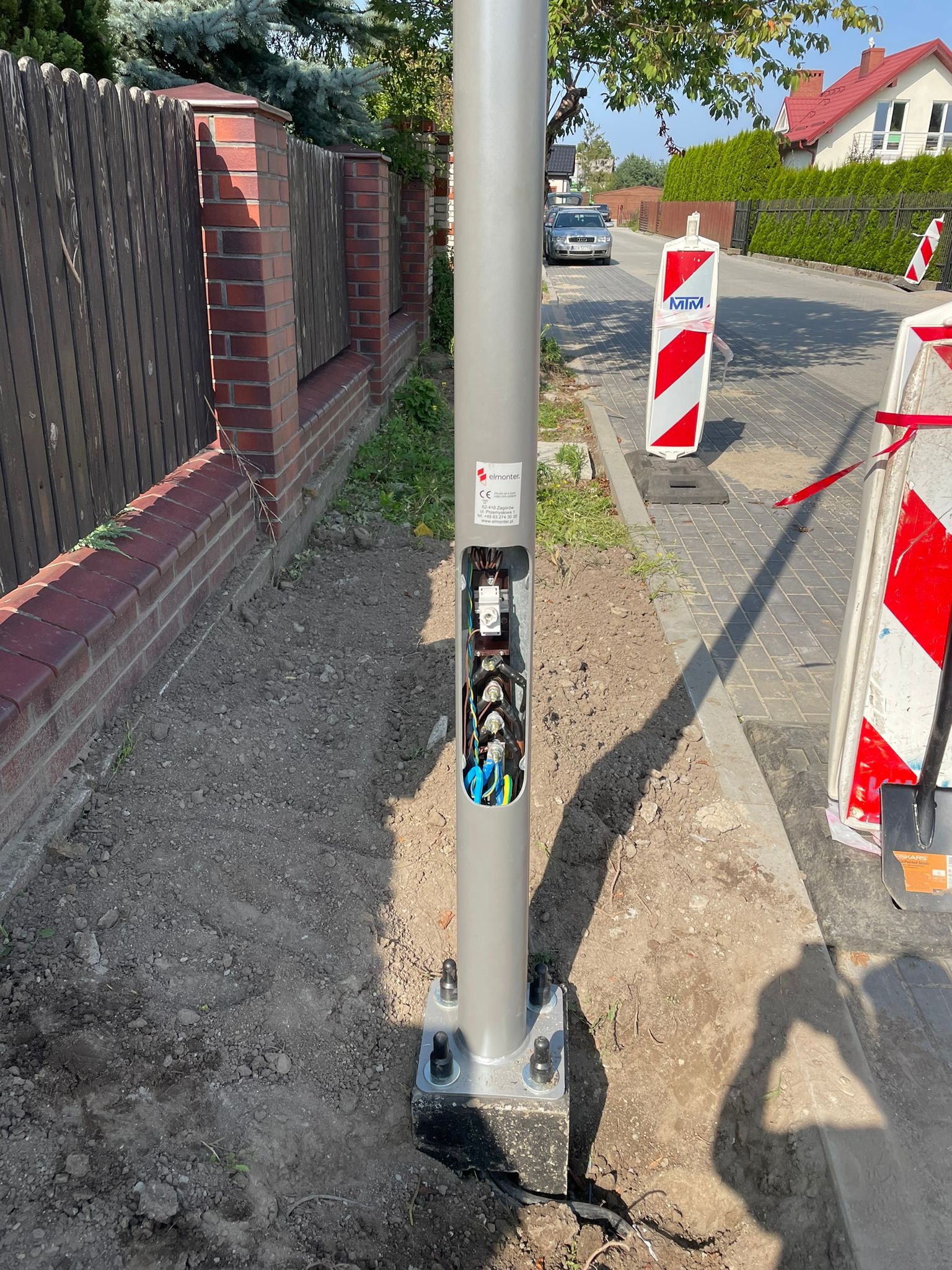 Otwarta skrzynka elektryczna w słupie oświetleniowym na zewnątrz, widoczne kable i złącza. Słup zamontowany na fundamencie, obok chodnik i ogrodzenie z cegły.